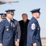 Man in a dark overcoat walking among uniformed military officers on a tarmac at an airfield