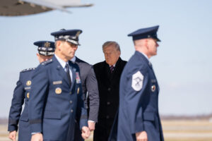 Man in a dark overcoat walking among uniformed military officers on a tarmac at an airfield