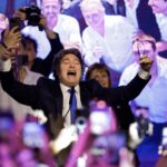 Man in a dark suit raises his arms and shouts onstage at a crowded event with cameras flashing behind him.