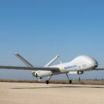 White Hermes 900 UAV on a runway with landing gear down, clear blue sky above.