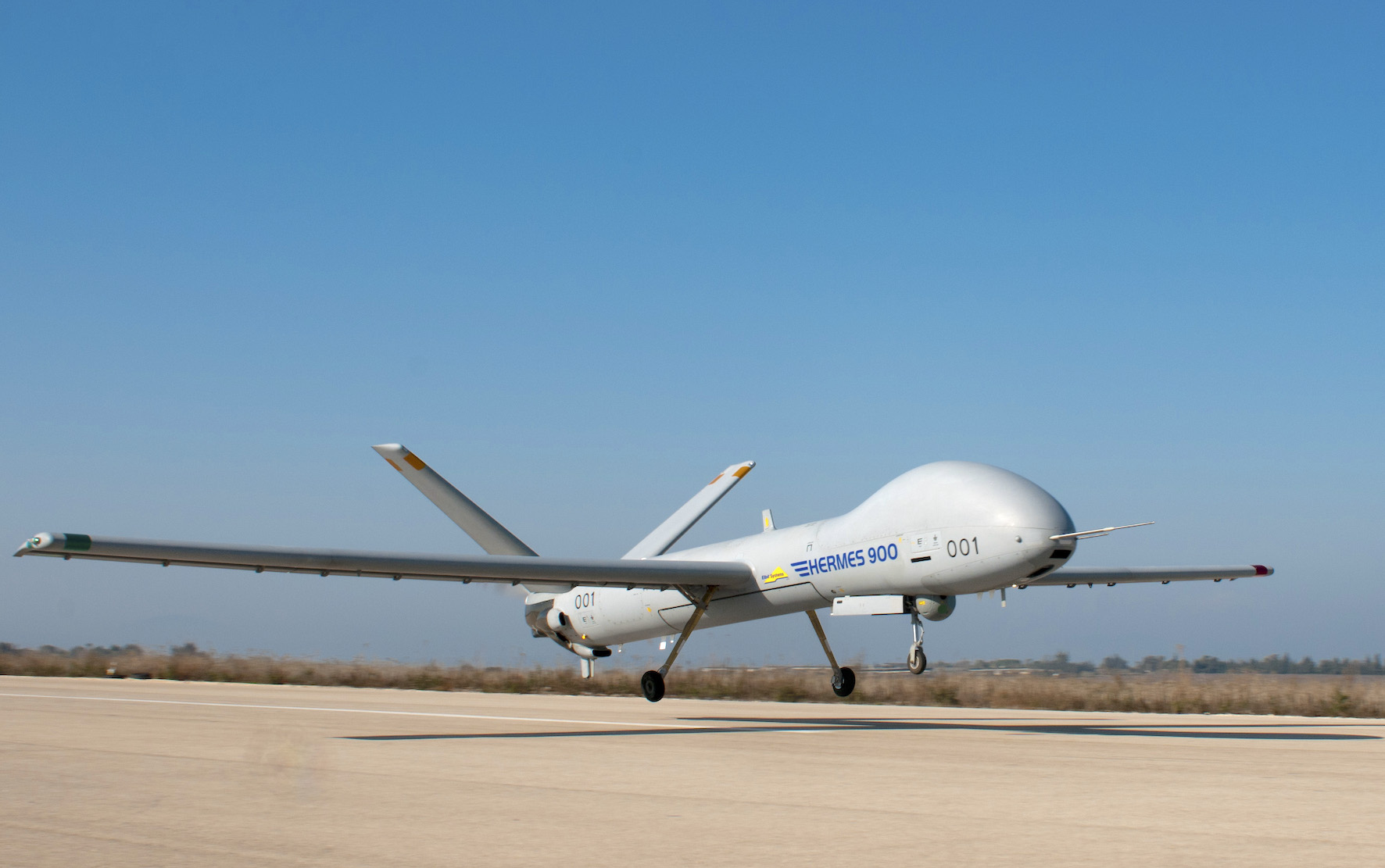 White Hermes 900 UAV on a runway with landing gear down, clear blue sky above.