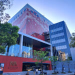 Modern glass-fronted building with a red patterned tower behind, trees along the sidewalk, and people entering.