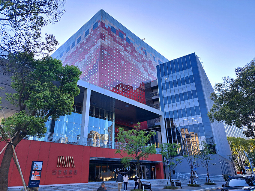 Modern glass-fronted building with a red patterned tower behind, trees along the sidewalk, and people entering.