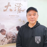 Smiling man in a black hoodie and cap posing beside a poster that reads 'A FOGGY TALE' with a map illustration.