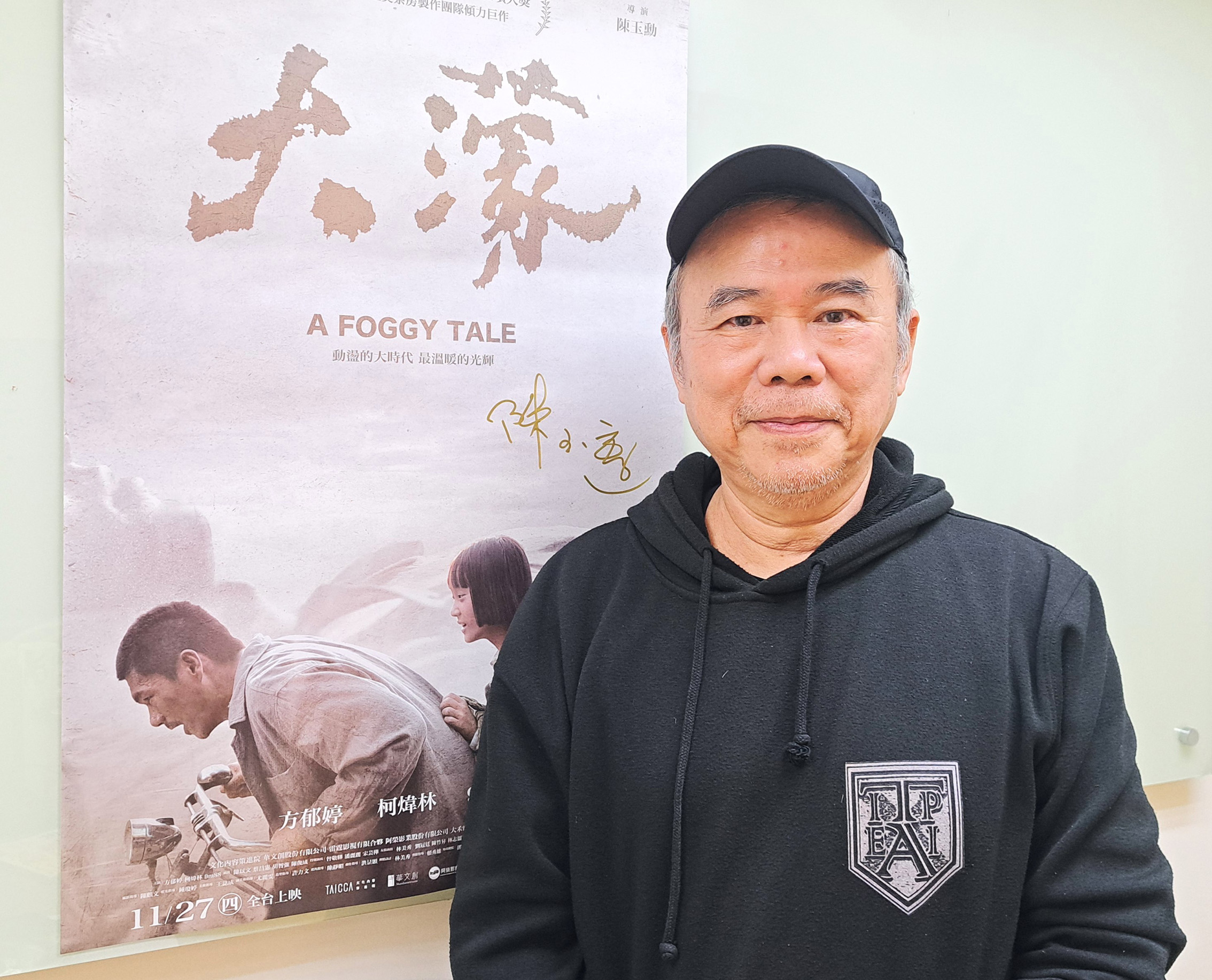 Smiling man in a black hoodie and cap posing beside a poster that reads 'A FOGGY TALE' with a map illustration.