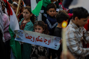 Young girl in a teal dress holds a banner with Arabic text at a crowded protest, surrounded by children and flags.