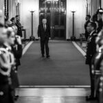 A man in a dark suit walks down a ceremonial hallway flanked by guards, with photographers in the foreground (black‑and‑white).