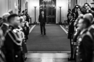 A man in a dark suit walks down a ceremonial hallway flanked by guards, with photographers in the foreground (black‑and‑white).