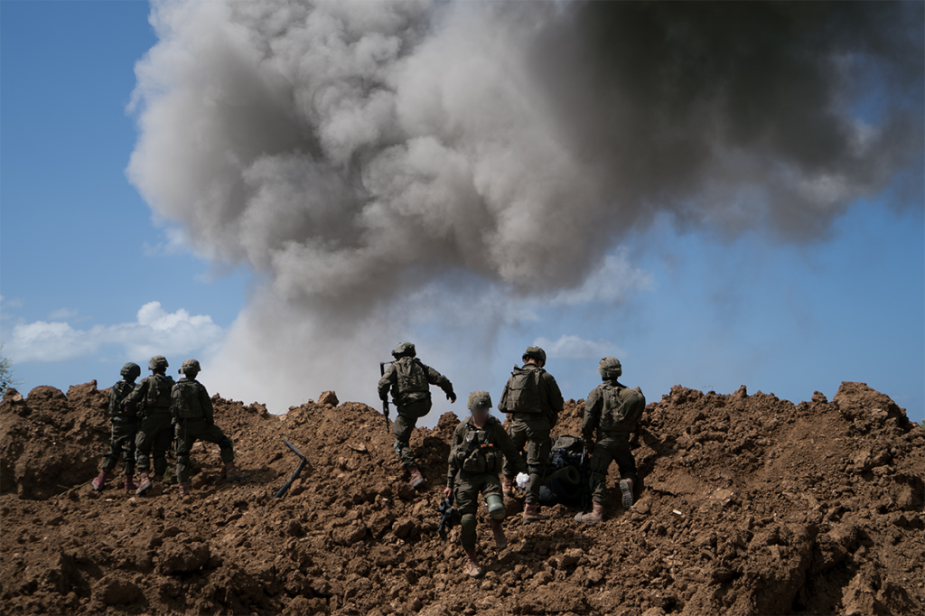 Soldiers in full combat gear ascend a muddy mound as a huge smoke plume rises behind them.