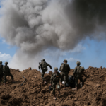Soldiers in full combat gear ascend a muddy mound as a huge smoke plume rises behind them.