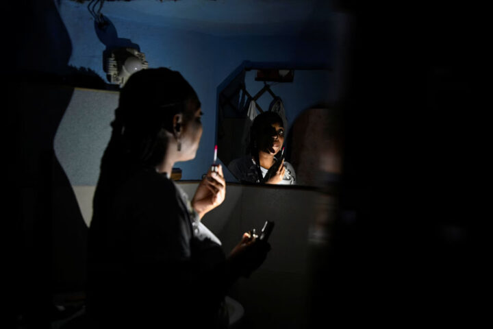 Woman applying lipstick while looking at her reflection in a mirror in a dimly lit room thought to be a dressing area