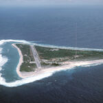 Aerial view of a small island with a straight runway, sandy beaches, and a tall radio antenna toward the coast.