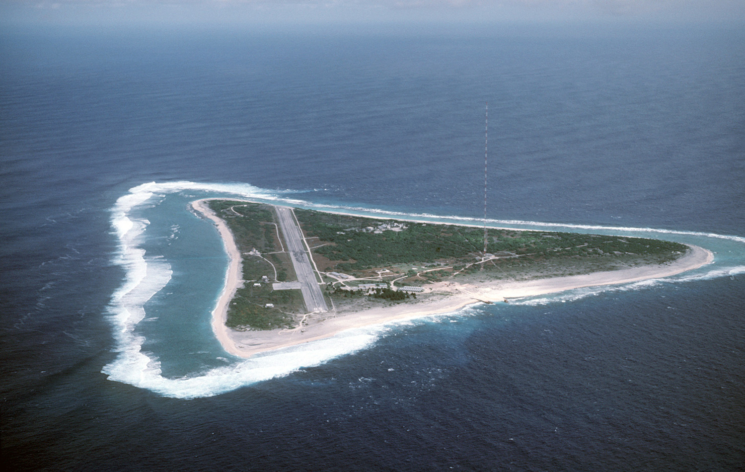 Aerial view of a small island with a straight runway, sandy beaches, and a tall radio antenna toward the coast.