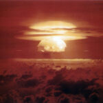 Nuclear explosion mushroom cloud rising above clouds at sunset over a dark horizon sea of clouds.