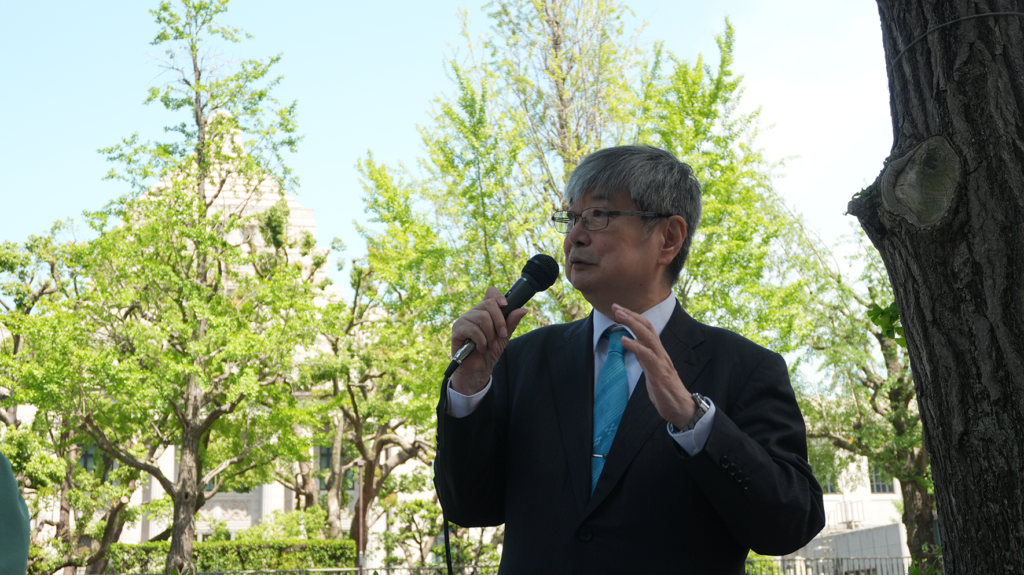 Man in a dark suit and blue tie speaks into a handheld microphone outdoors, with green trees in the background.
