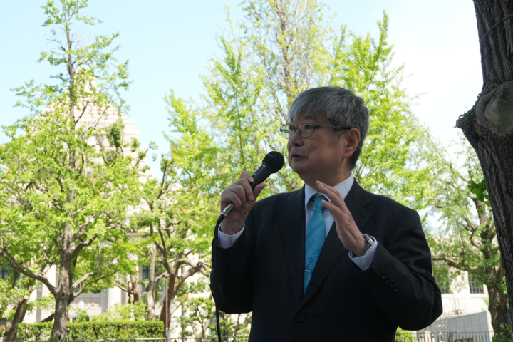 Man in a dark suit and blue tie speaks into a handheld microphone outdoors, with green trees in the background.