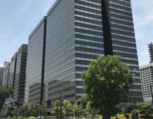 Tall modern office building with horizontal glass bands, blue sky, and green trees in foreground.