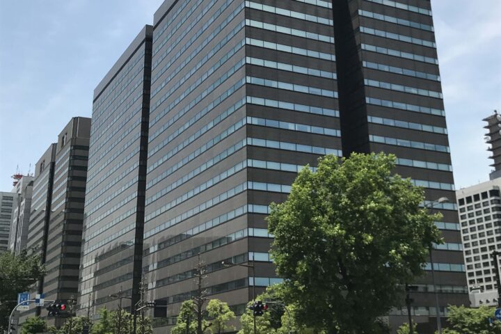 Tall modern office building with horizontal glass bands, blue sky, and green trees in foreground.