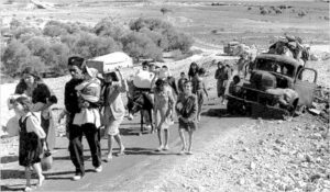 Group of civilians walking along a dusty road carrying belongings, with a burned-out car and desert landscape in the background (historical scene).
