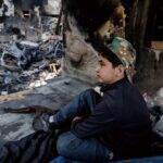 Young boy in a camouflage cap sits among charred debris of a ruined building, gazing to the right.