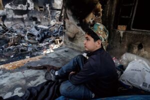 Young boy in a camouflage cap sits among charred debris of a ruined building, gazing to the right.