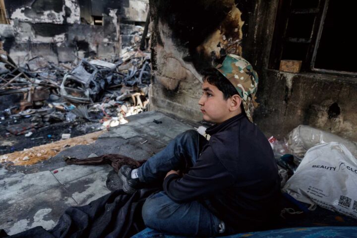 Young boy in a camouflage cap sits among charred debris of a ruined building, gazing to the right.