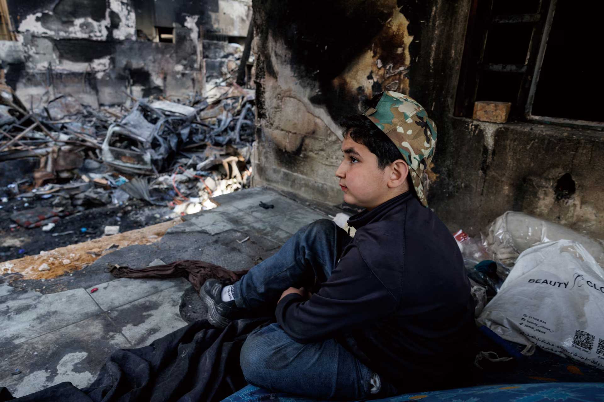 Young boy in a camouflage cap sits among charred debris of a ruined building, gazing to the right.
