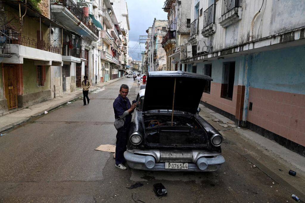 Mechanic in overalls standing by a vintage car with its hood open on a narrow, rundown street lined with balconies and weathered buildings.