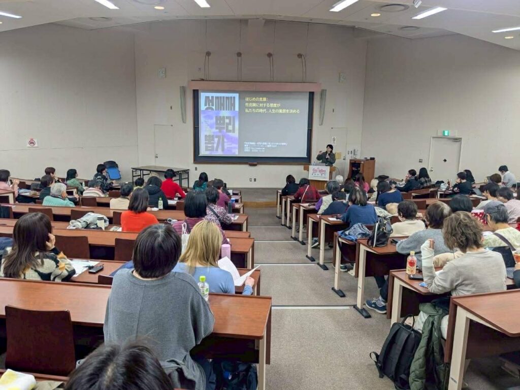 Lecture hall with a speaker at a podium presenting to an audience in tiered rows, projected slide visible.
