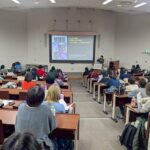 Lecture hall with a speaker at a podium presenting to an audience in tiered rows, projected slide visible.