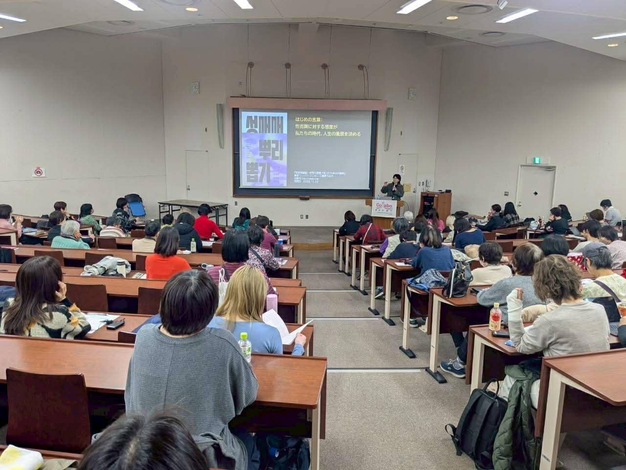 Lecture hall with a speaker at a podium presenting to an audience in tiered rows, projected slide visible.