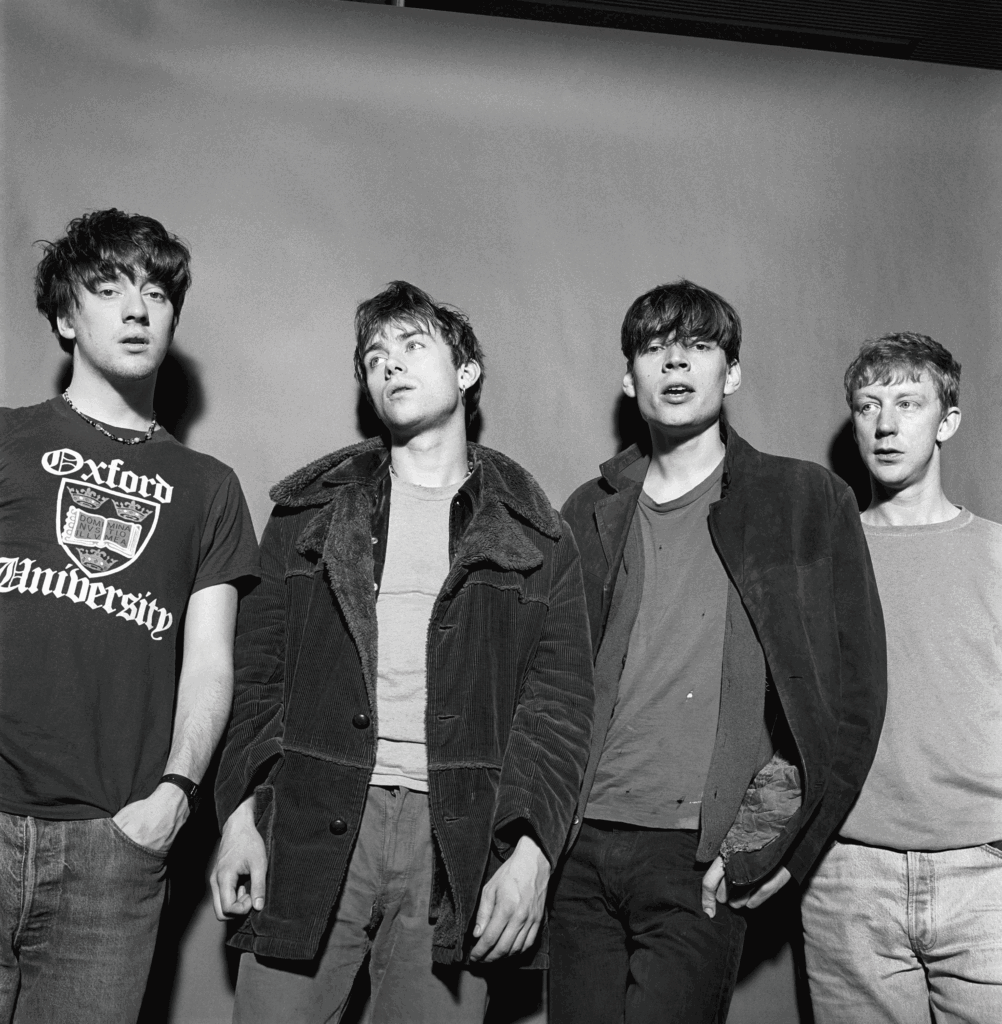 Four young men stand together shoulder to shoulder in casual outfits, black‑and‑white photo.