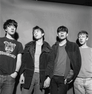 Four young men stand together shoulder to shoulder in casual outfits, black‑and‑white photo.