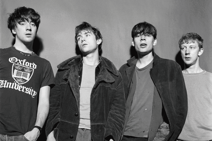 Four young men stand together shoulder to shoulder in casual outfits, black‑and‑white photo.