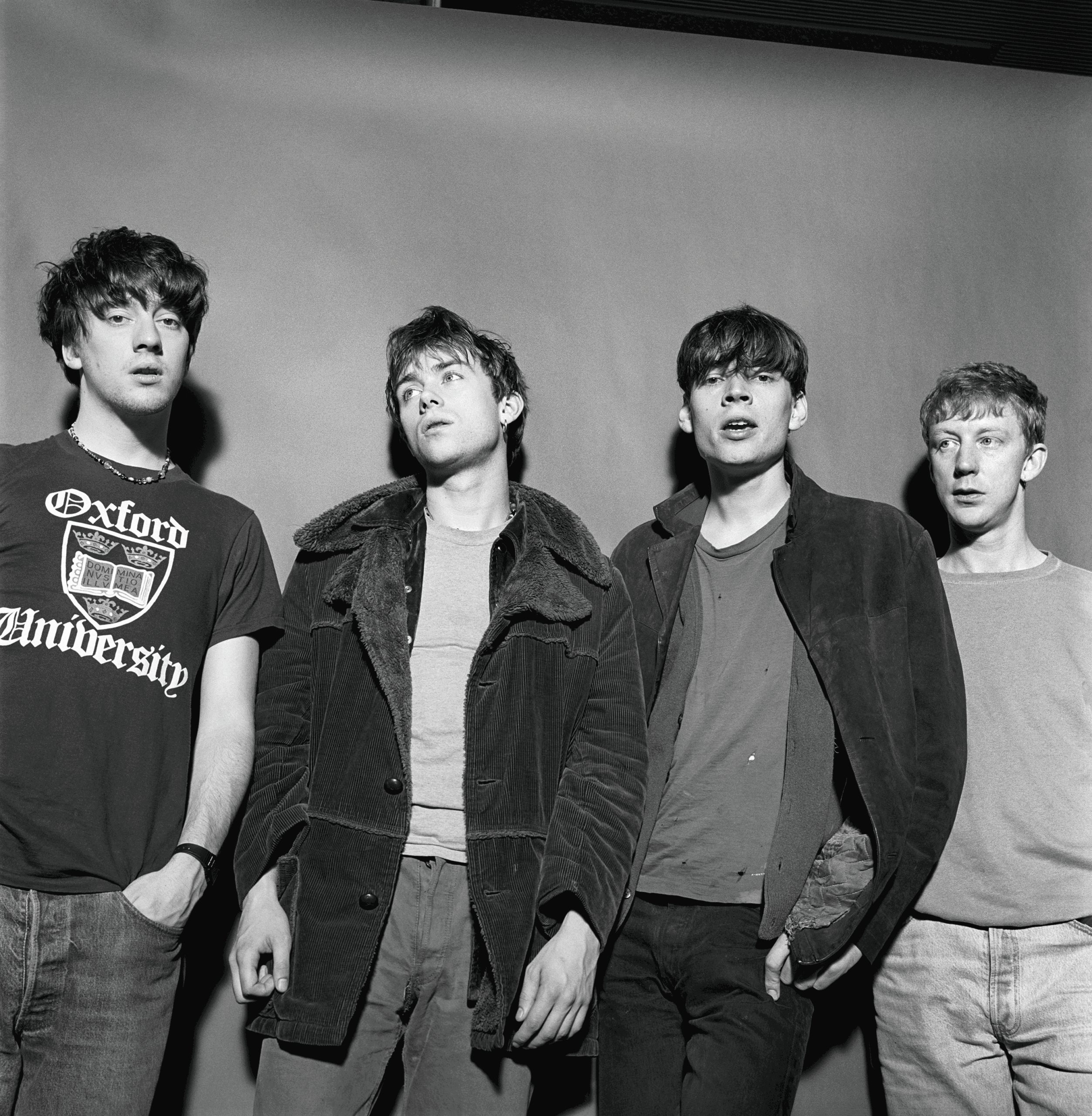 Four young men stand together shoulder to shoulder in casual outfits, black‑and‑white photo.
