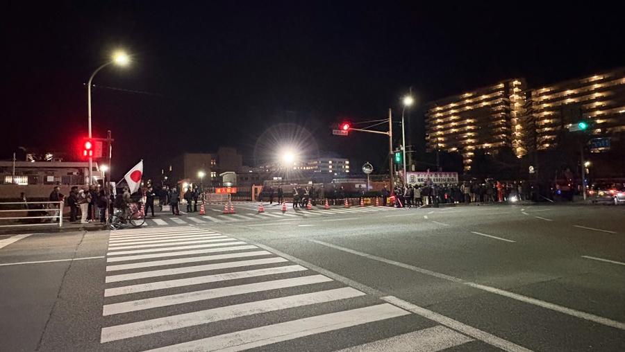 Nighttime city intersection with a crowd along the crosswalk, a Japanese flag, traffic cones, and barricades under streetlights.
