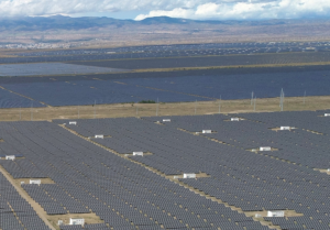 A vast solar power farm with rows of dark solar panels across flat brown terrain, distant mountains on the horizon and utility poles in the background.