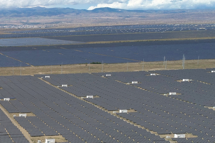 A vast solar power farm with rows of dark solar panels across flat brown terrain, distant mountains on the horizon and utility poles in the background.