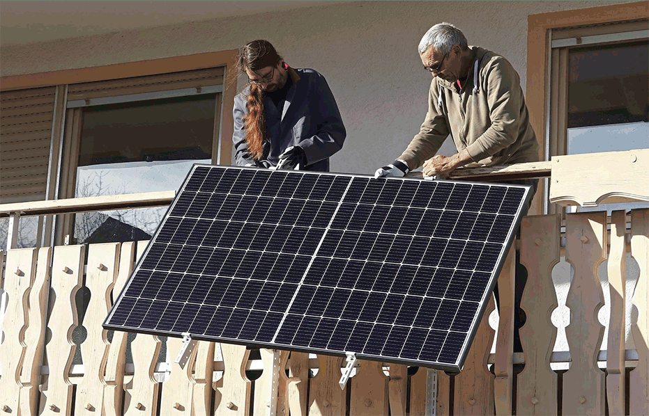 Two people install a solar panel on a balcony railing of a house, mounting it for solar power.
