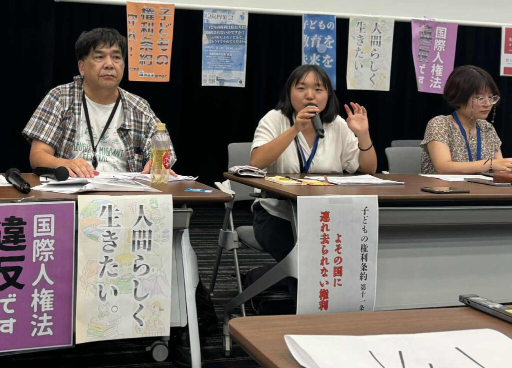 Three panelists sit at a long table; the middle person speaks into a microphone during a discussion or protest event, with banners behind them.