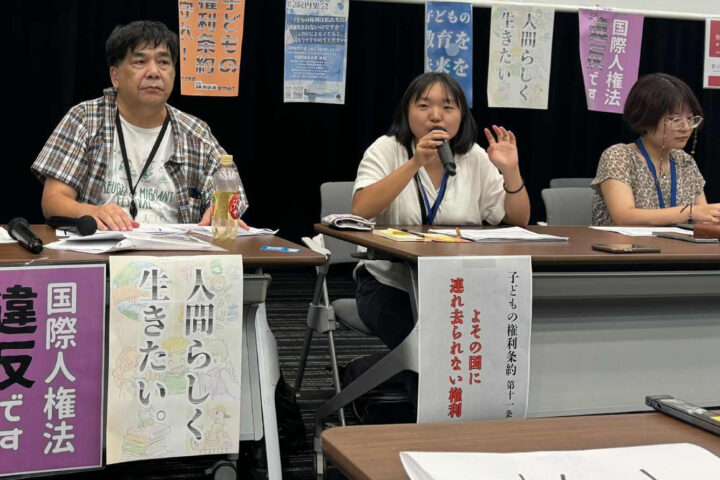 Three panelists sit at a long table; the middle person speaks into a microphone during a discussion or protest event, with banners behind them.