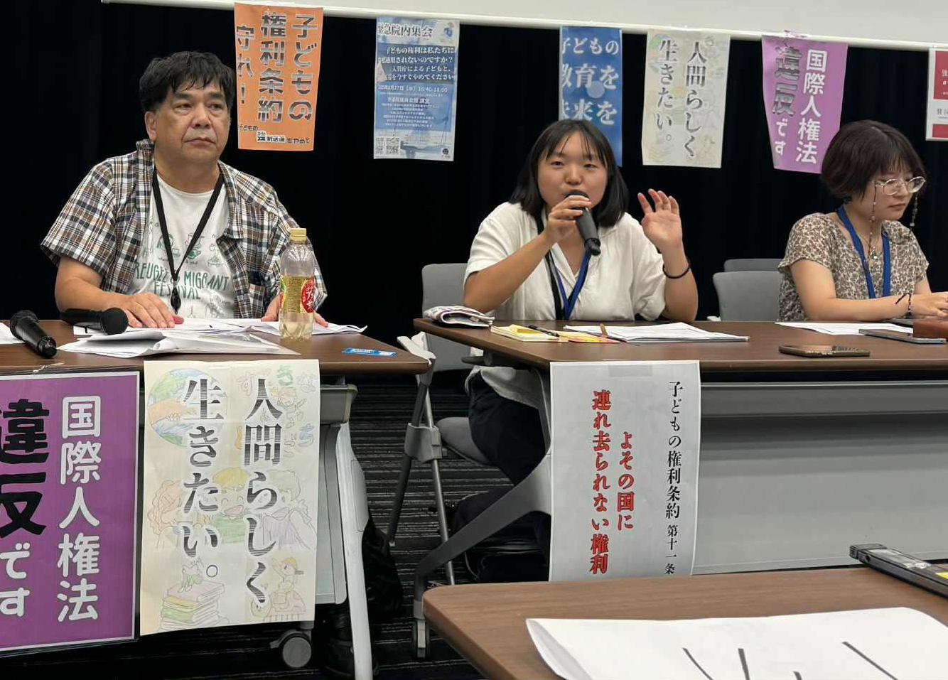Three panelists sit at a long table; the middle person speaks into a microphone during a discussion or protest event, with banners behind them.