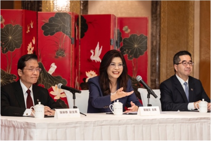 Panel discussion with three speakers seated at a long table, microphones in front, and a decorative red screen behind them.