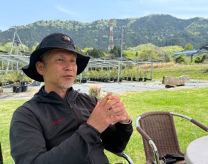 Man in a black jacket and bucket hat sits at an outdoor table with mountains in the background, suggesting a relaxed outdoor setting.