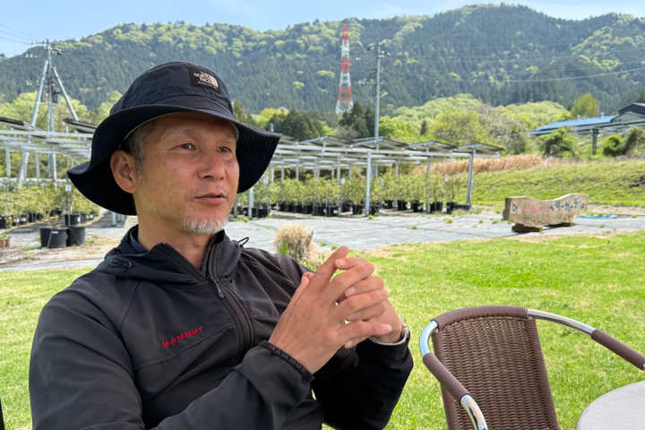 Man in a black jacket and bucket hat sits at an outdoor table with mountains in the background, suggesting a relaxed outdoor setting.