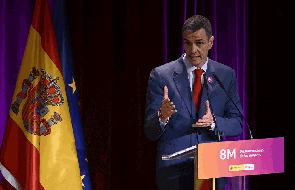 Man in a blue suit speaks at a podium for International Women's Day (8M), with Spain and EU flags behind him.