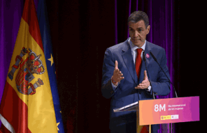 Man in a blue suit speaks at a podium for International Women's Day (8M), with Spain and EU flags behind him.