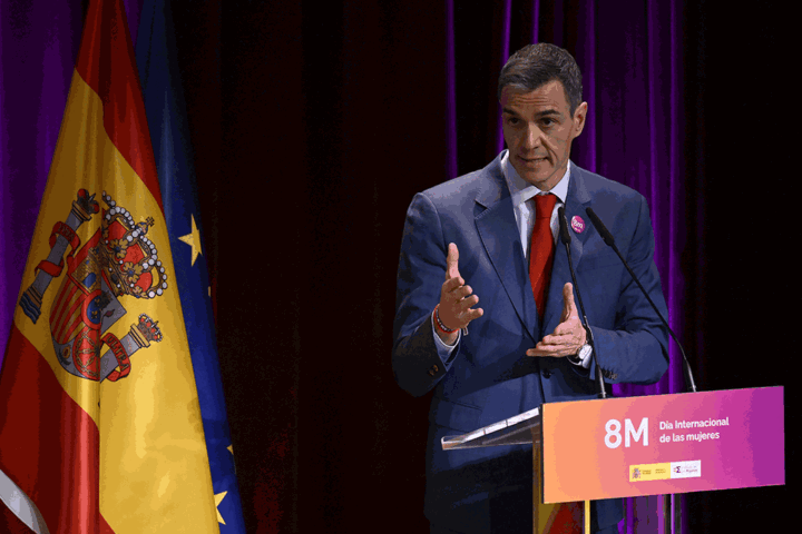 Man in a blue suit speaks at a podium for International Women's Day (8M), with Spain and EU flags behind him.