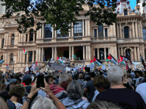 Large crowd of protesters in front of a historic sandstone building, waving Palestinian flags and holding signs.