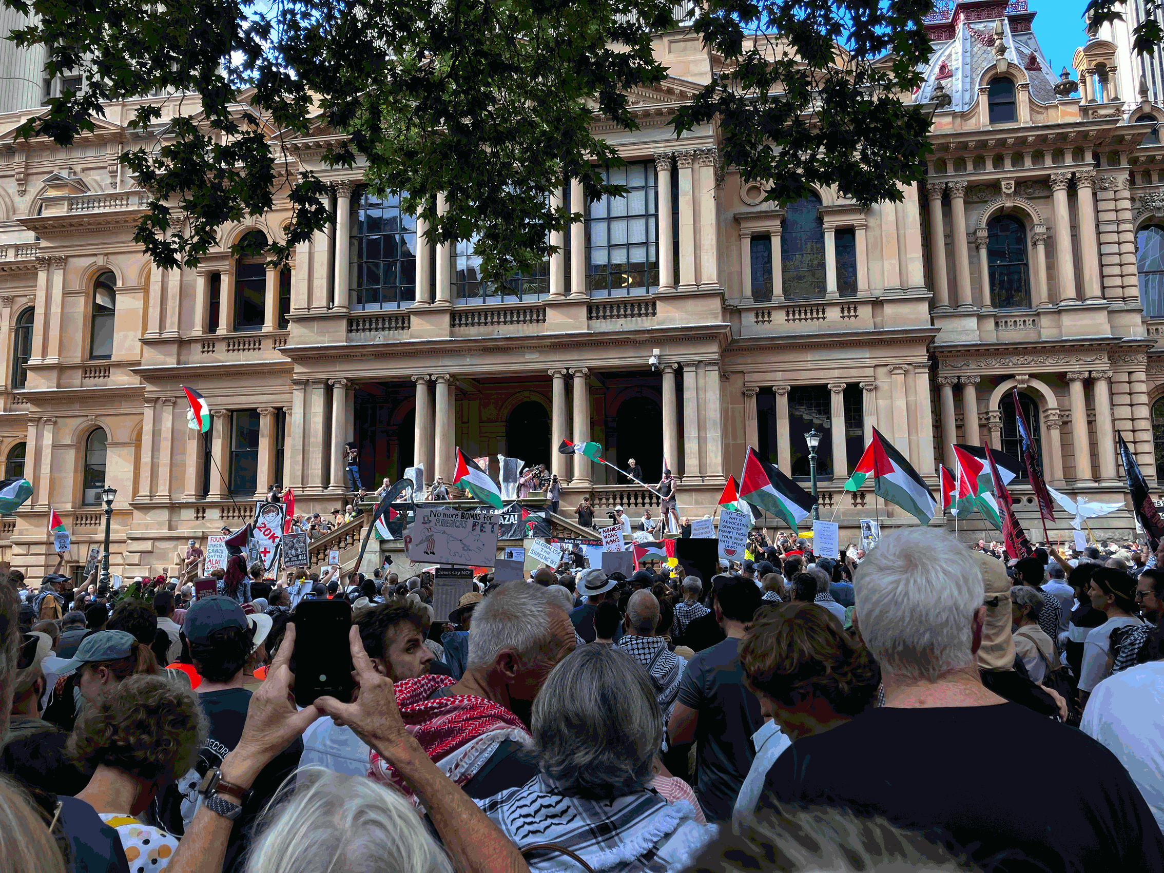 Large crowd of protesters in front of a historic sandstone building, waving Palestinian flags and holding signs.